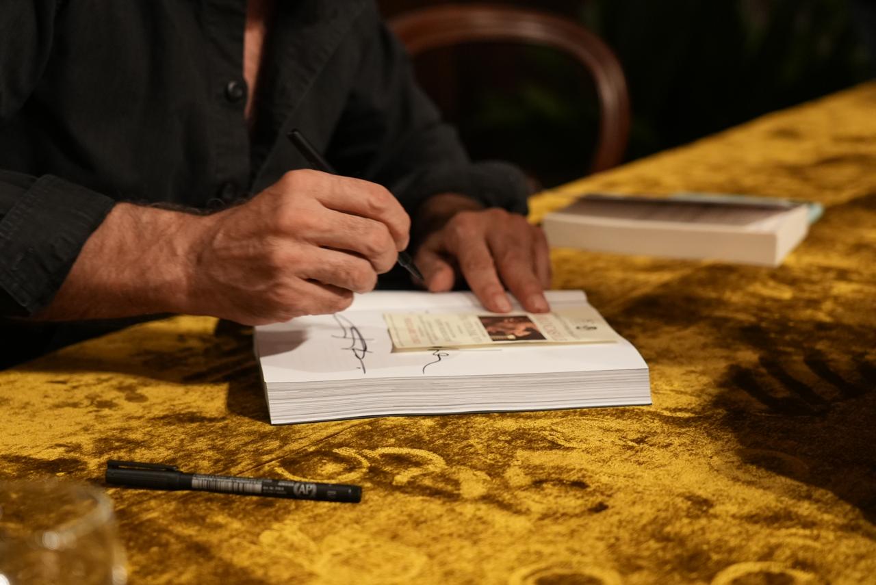 Una mano firma il frontespizio del libro, un pennarello sul tavolo